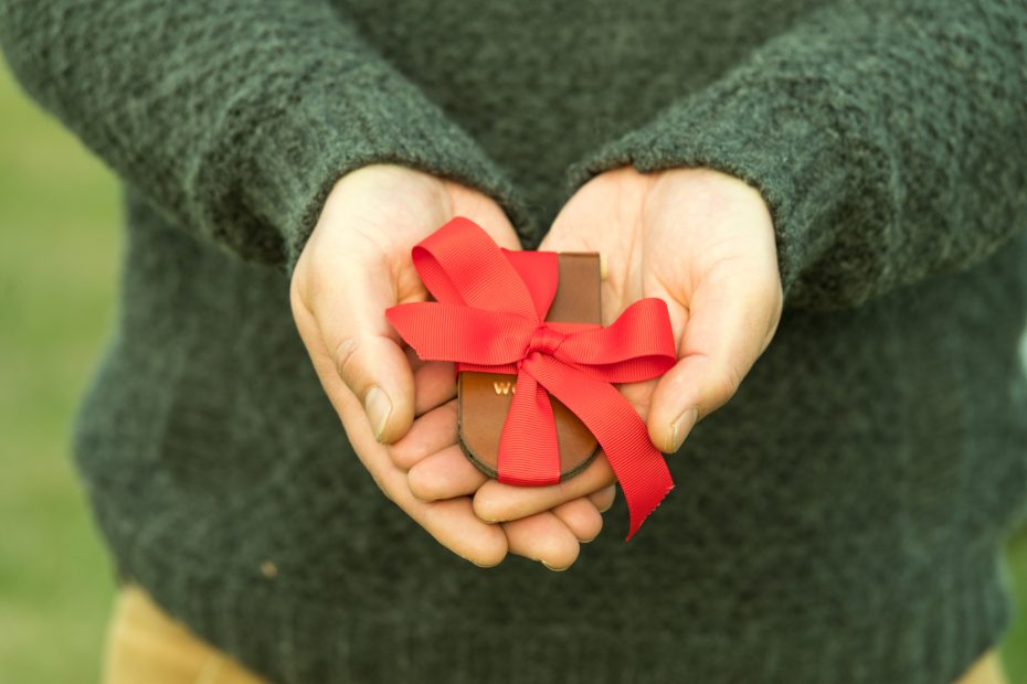 Man in a green wool sweater holding a small gift wrapped in a red ribbon in his hands. His hands make a heart shape from the palms meeting and the fingers overdressed.