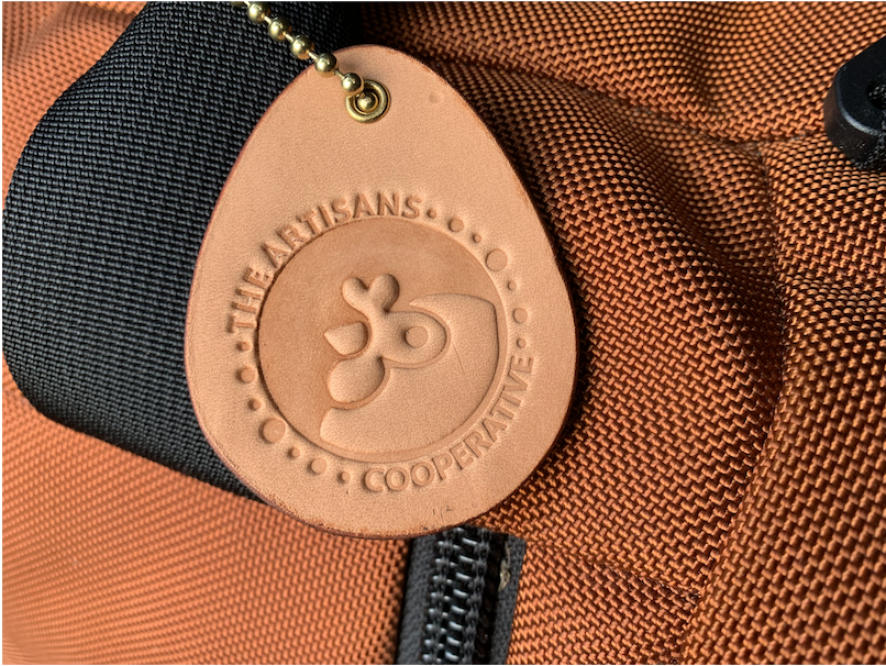 Leather Luggage Tag with the Artisans Cooperative Logo debossed into the leather (stamped in). Has a brass ball chain and is attached to an orange luggage bag.