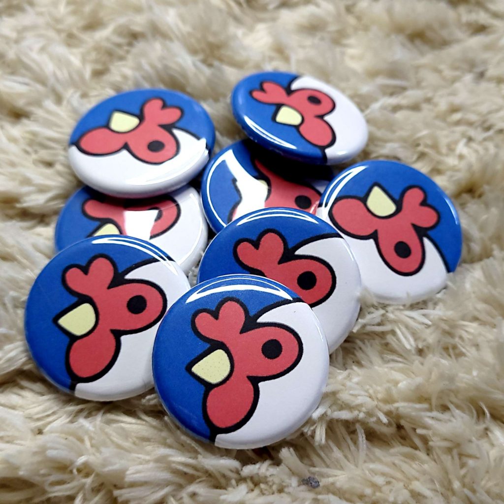 A pile of 1.25" Finback Buttons with the Artisans Cooperative Chicken Mascot in a Woman's Hand Against a Fabric Backdrop for an Artist Fundraiser on a Furry Rug