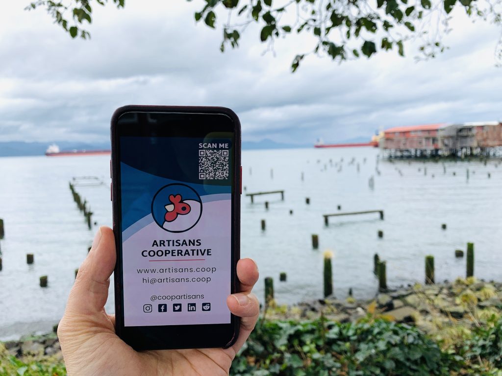 A woman holds up a phone displaying the Artisans Cooperative digital business card on a riverfront