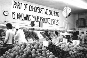 Historic photo of Black American shoppers at a cooperative grocery store in the product section. Above the product hangs a sign that says "Part of Co-operative Shopping is Knowing Your Produce Prices." 
