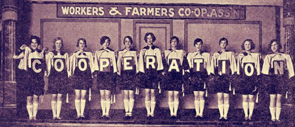 Historic photo of a group of women lined up underneath a sign for the Workers & Farmers Co-op Association. Each woman is holding one letter in the word COOPERATION so together they spell the entire word. Era is likely 1900-1930.