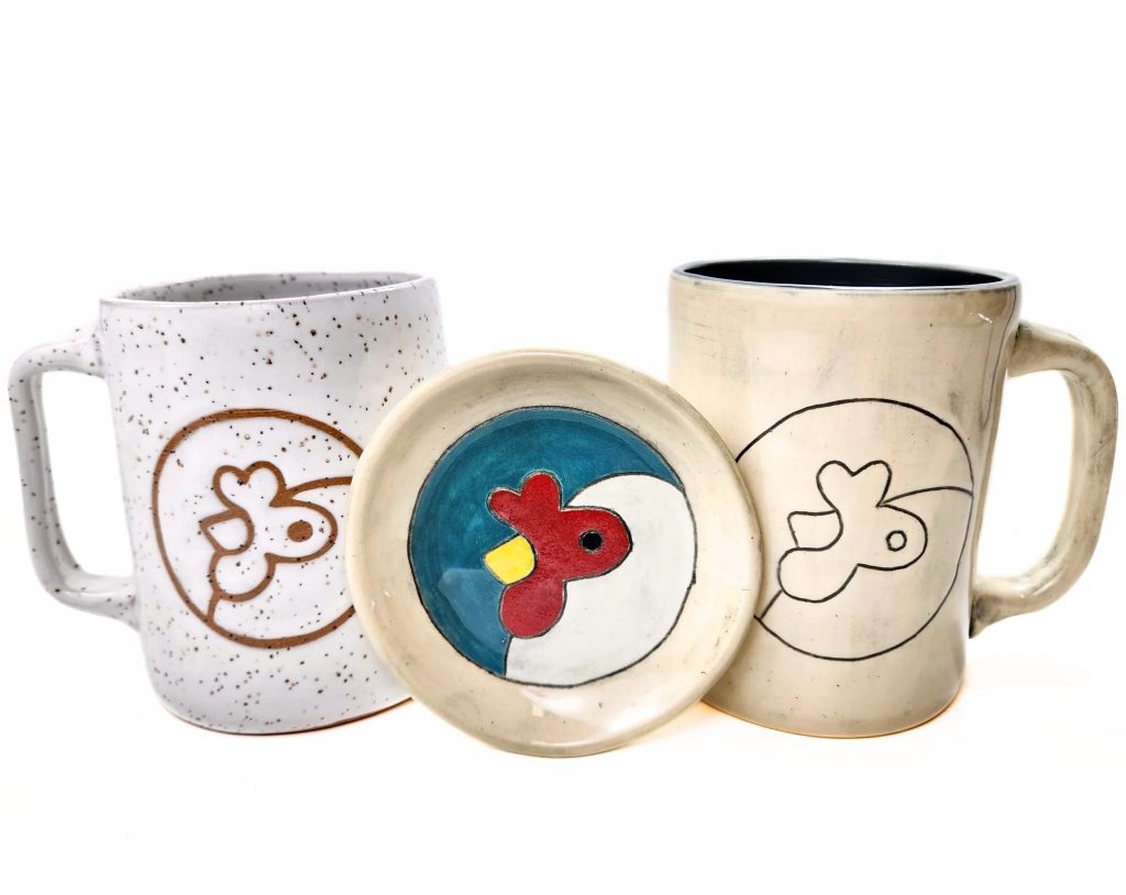 Two handmade pottery mugs made from stoneware. One mug is white speckled and the other is an off-white with a black line glaze. The dish is full color painted glaze. All feature the Artisans Cooperative logo chicken mascot