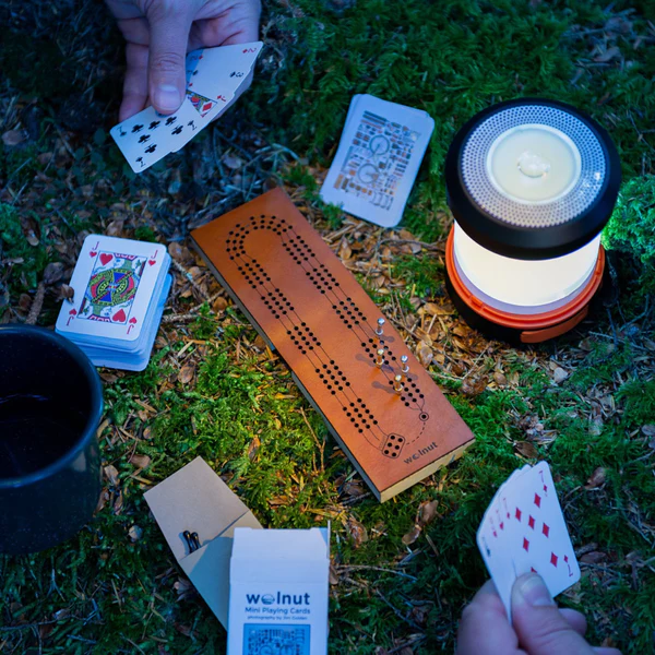 An evening photo of a mini cribbage board sitting in a patch of moss. Next to it is a lantern lighting the area while the hands of two people can be seen playing across from each other, each holding a mini spread of playing cards.