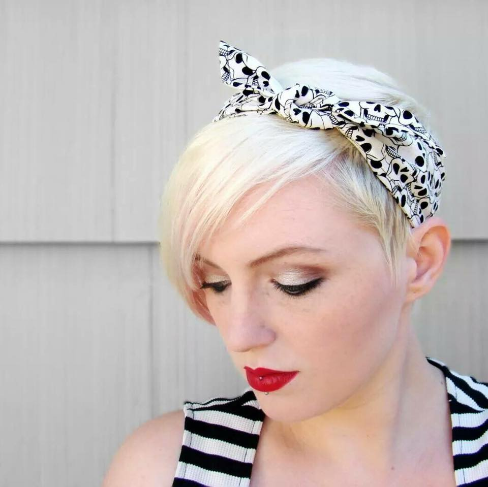 A photo of a 3/4 view of a blonde woman's head as she looks to the bottom left corner. In her pixie cut style hair is a black and white fabric headband with a skull pattern. The headband is tied in a knot and slightly offset from the top of her head. 