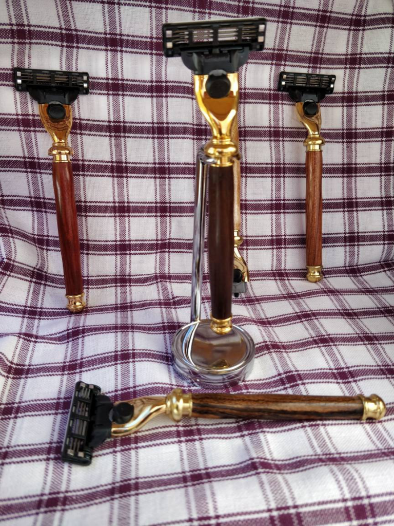 A photo of four shaving razors on a red and white plaid fabric background. The razors all have brass hardware and have handles made from various shades of smooth wood. One razor lays flat while the others are positioned upright.
