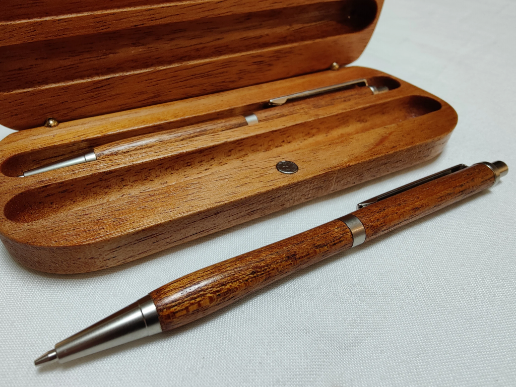 A photo of a silver pen with a polished wooden body. Behind the pen is a magnetic wooden pen case flipped open. The case has two slots, the rear of which is holding a matching pen to the one in front of the case. 