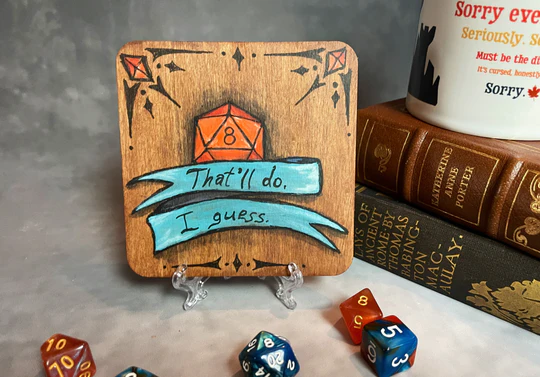 A wooden coaster with a painting of an orange d20 die reading a number 8. The die is overlaid with a blue ribbon that reads, "That'll do. I guess."