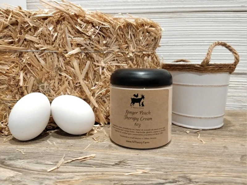 A photo of a cylindrical container of body cream placed between a pair of white eggs on the left and a white, metal bucket on the right, all sitting in front of small hay bale that continues out of left frame.