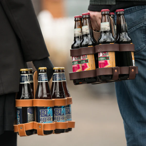 A closeup photo of two six packs of beer bottles in leather carrying cases, both carried by a person on either side of frame. The case on the left is made from  light brown leather straps , while the case on the right is made from dark brown leather straps. 