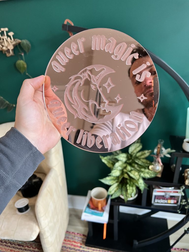 A hand holding out a circular, rose-gold mirror that reads "queer magic is queer liberation." Surrounded by the text is a horse head in front of a crescent moon and stars.
