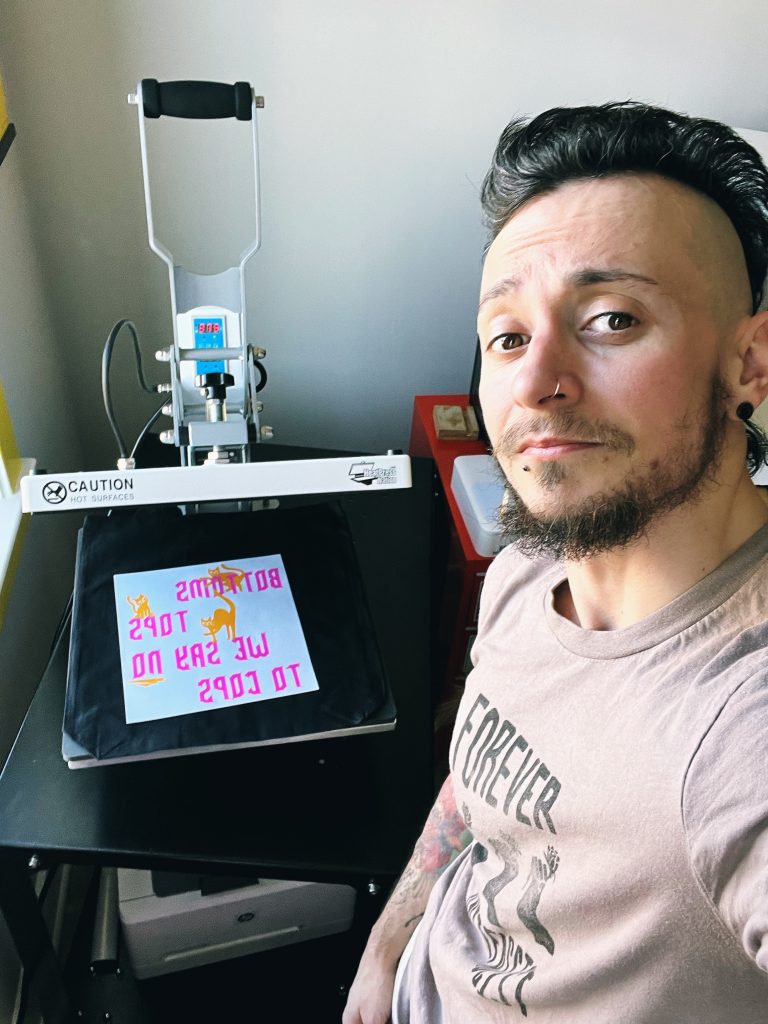 A selfie of a person (Raffi) showing off their workstation with one of their pieces inside a heat printing press. In pink, mirrored text, the art reads, "Bottoms Tops We say no to cops."