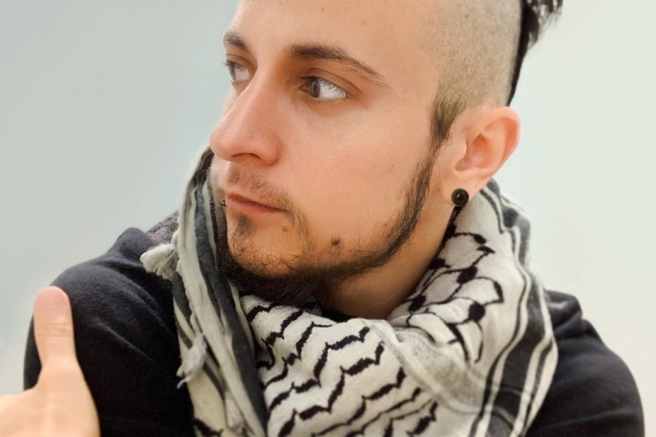 A photo of Raffi, a person with short brown hair with shaved sides, and a short beard. They're wearing a black long sleeve shirt, a white and black-striped triangle scarf, and a beaded bracelet.