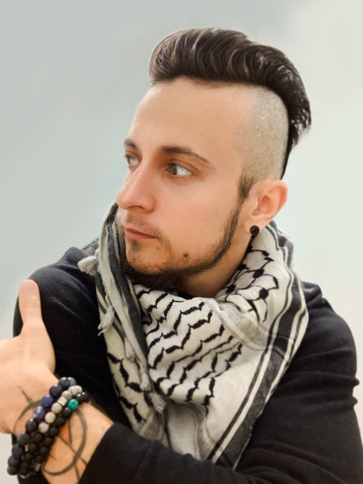 A photo of Raffi, a person with short brown hair with shaved sides, and a short beard. They're wearing a black long sleeve shirt, a white and black-striped triangle scarf, and a beaded bracelet.