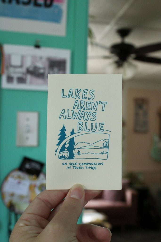 Picture of hand holding mini zine titled "Lakes aren't always blue: on self compassion in tough times".