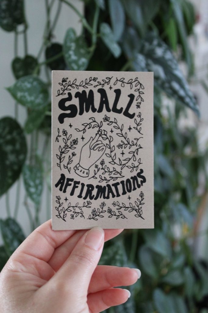 Picture of hand holding mini zine titled "Small Affirmations" in front of a leafy green background.