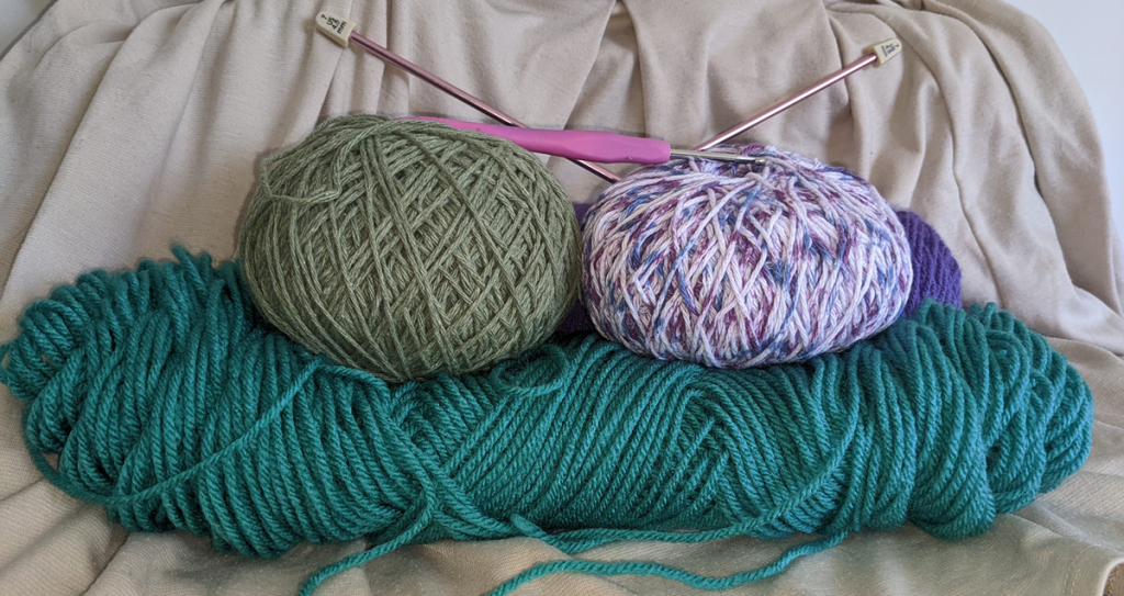 A close up of three different balls of yarn- one of pale olive green, one of multicolor shades of light purple, and an oblong one of medium teal. 