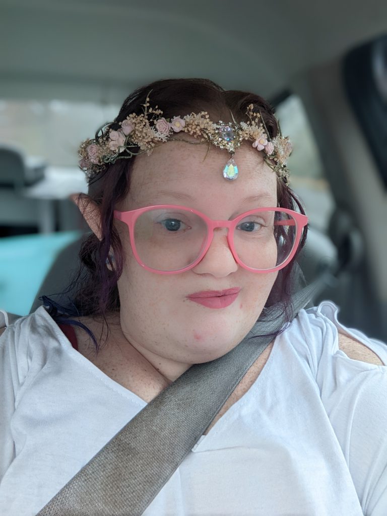 A photo of Elizabeth (Lizzy) sitting in a car. She has brown hair and is wearing a white shirt, as well as pink glasses and pink lipstick. She's also wearing a elf ears and a floral crown with pale pink flowers and a white teardrop gem dangling from the middle.
