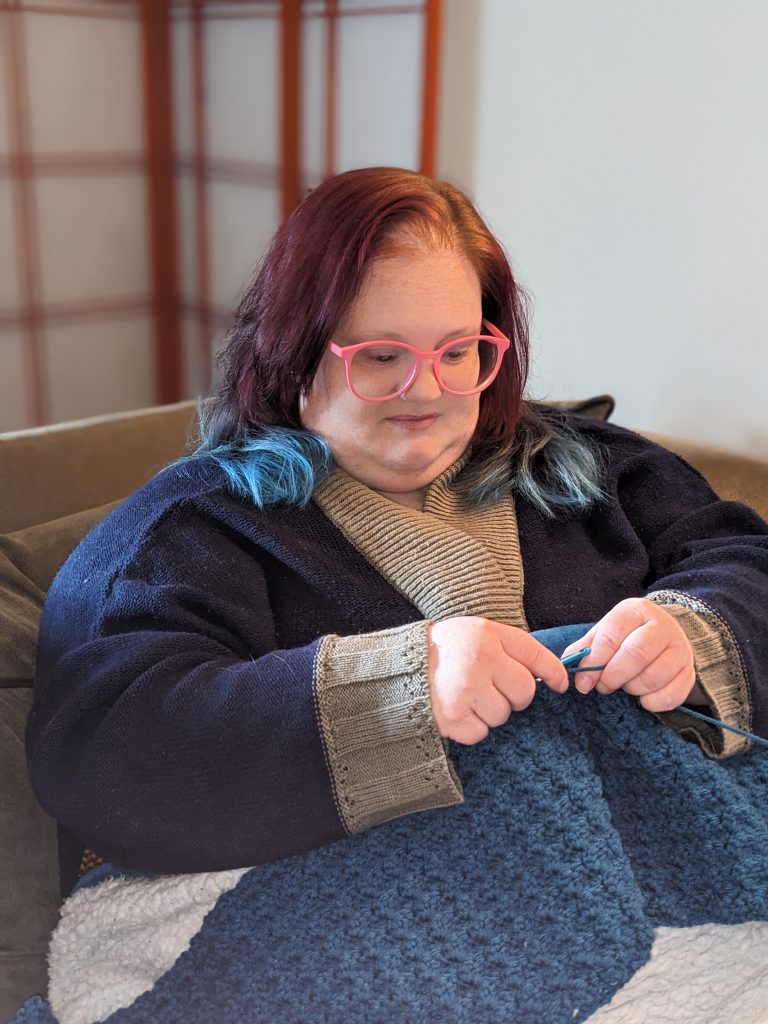 A photo of Lizzy sitting on a couch while working on a piece of embroidery made with blue yarn.