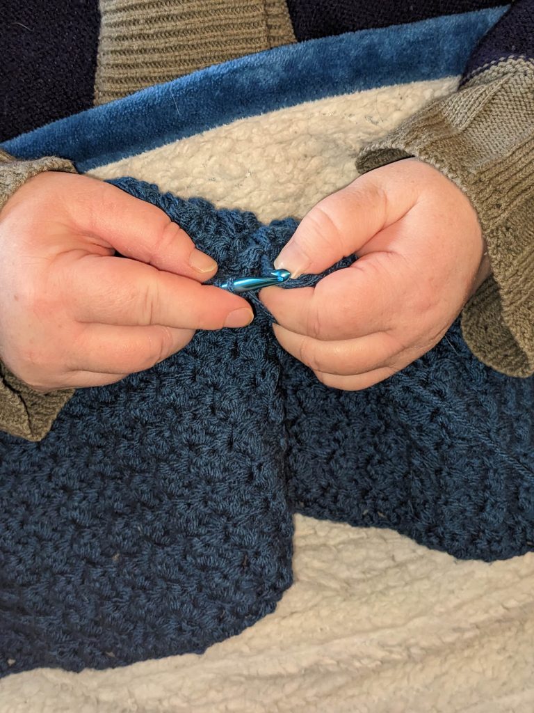A close-up photo of Lizzy's hands while she works on a piece of embroidery made with blue yarn. 