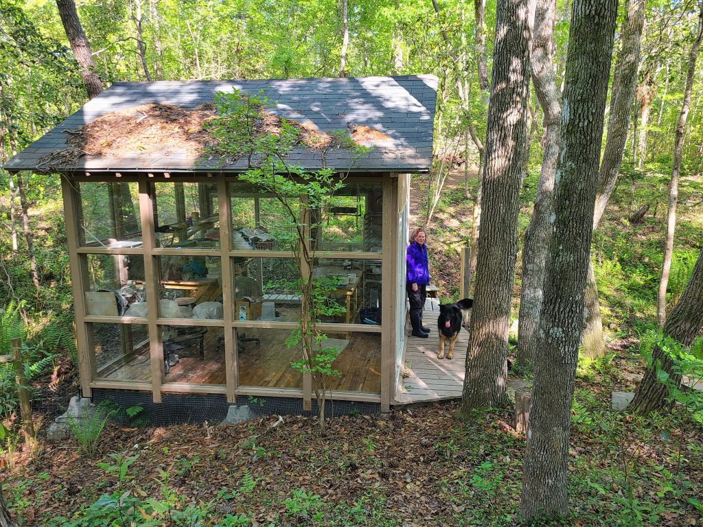 A photo of Erin standing with her dog at the edge of her work shed, which is surrounded by forest. 
