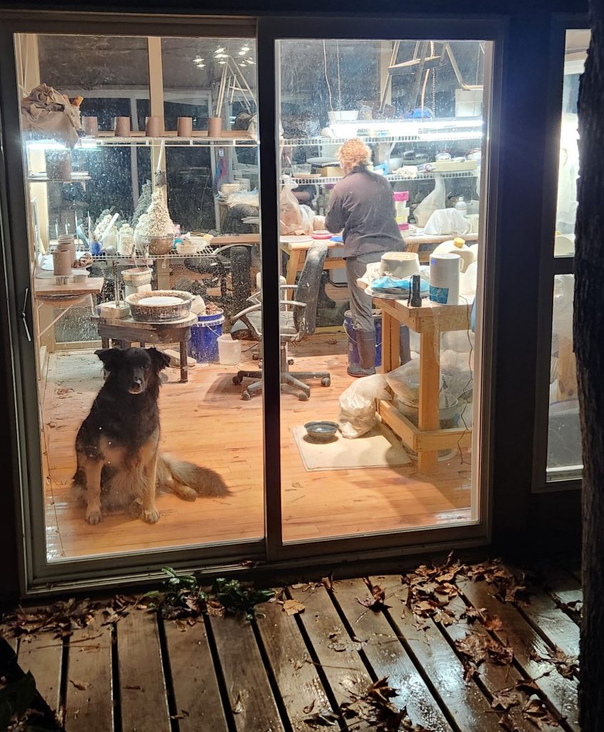 A nighttime photo from outside of Erin's workshop while she works with her back turned to the camera. Her dog stares out from the workshop at the viewer. 