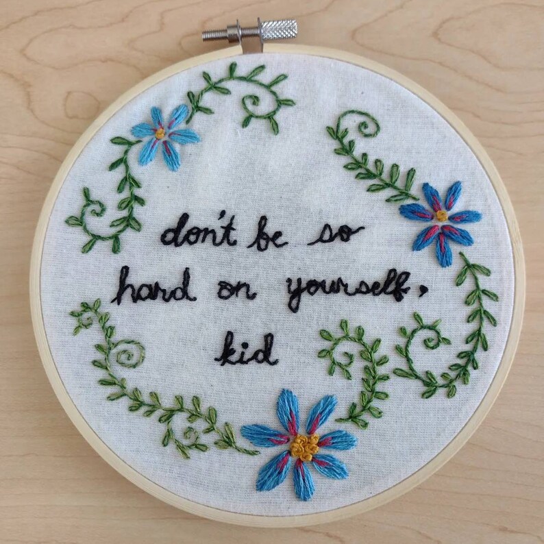 Photo of a hand embroidered artwork. There are blue flowers and swirling leaves surrounding the text in the center of the circular work. The text reads "don't be so hard on yourself, kid".