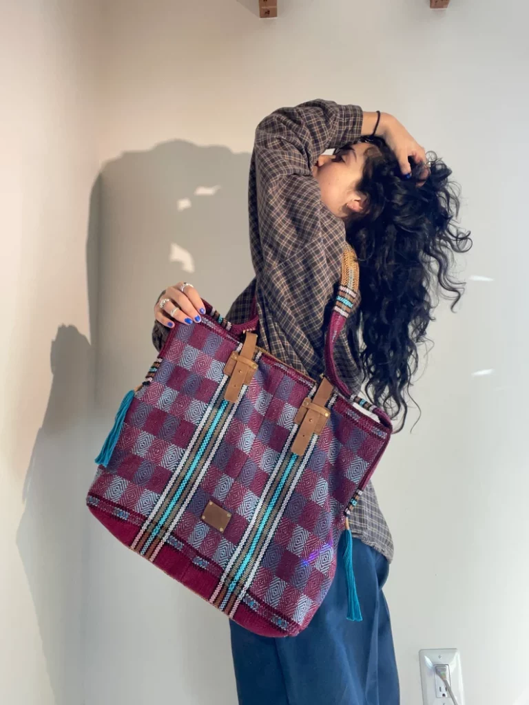 Photo of a woman holding a tote bag. The woman is standing profile to the camera, tote bag proudly on her shoulder. The woman is running her hand through her hair. The tote bag on her shoulder is beautifully hand woven. It has a checkerboard design, with a variety of purple and blue checks. 