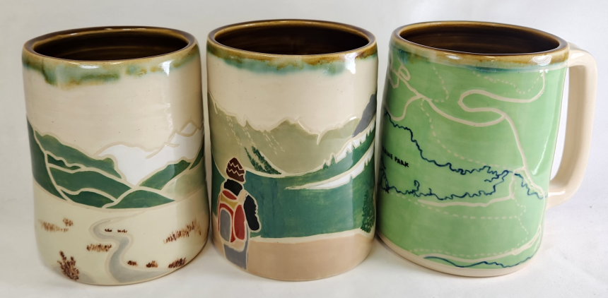 A picture of three handmade mugs. One has a beautiful green map painted on it, and the other two have landscapes painted on them.