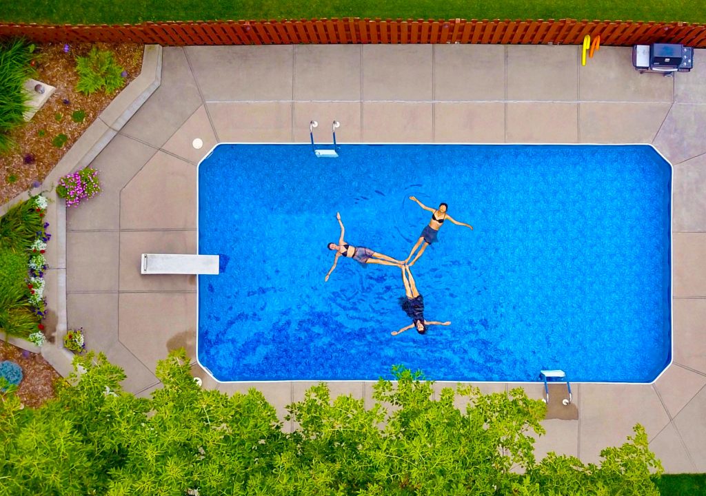 Aerial shot of a swimming pool as seen from the air like a bird's eye view. In the pool are three women floating on the surface with their arms outstretched and their feet linked together to form a trifecta or triad shape. Photo by Bruce Christianson for Unsplash