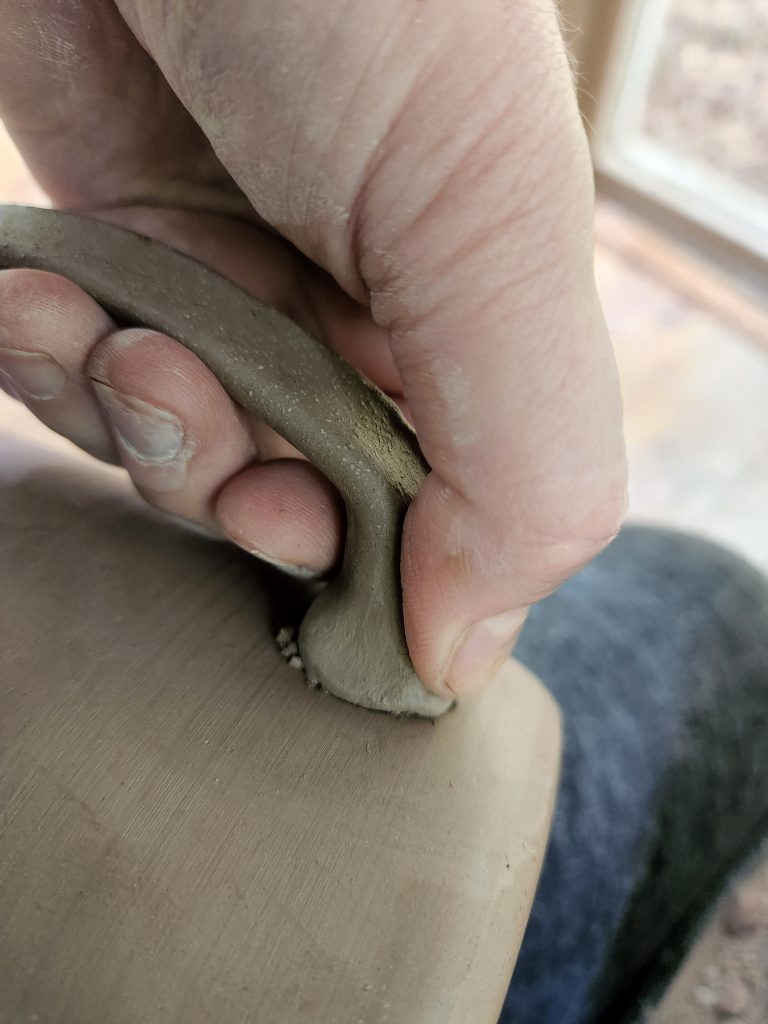 A photo of Erin's hand attaching a handle to an unfired pottery mug. 
