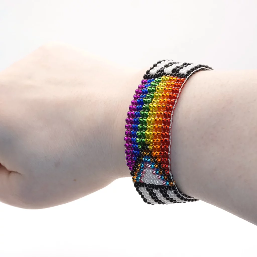On a wrist sits a bracelet made of small beads woven together. The bands are long black and white stripes while the center beads are arranged in the progressive pride flag colors. 