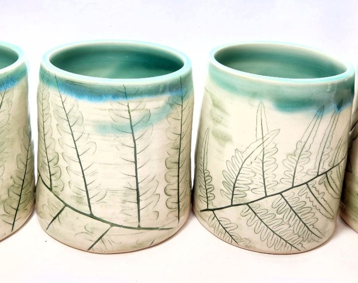 A photo of two off-white mugs with teal rims and fern impressions on the sides.