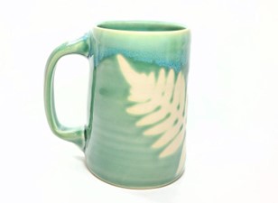 A photo of a light green mug with a yellowish/off-white fern impression on the side.