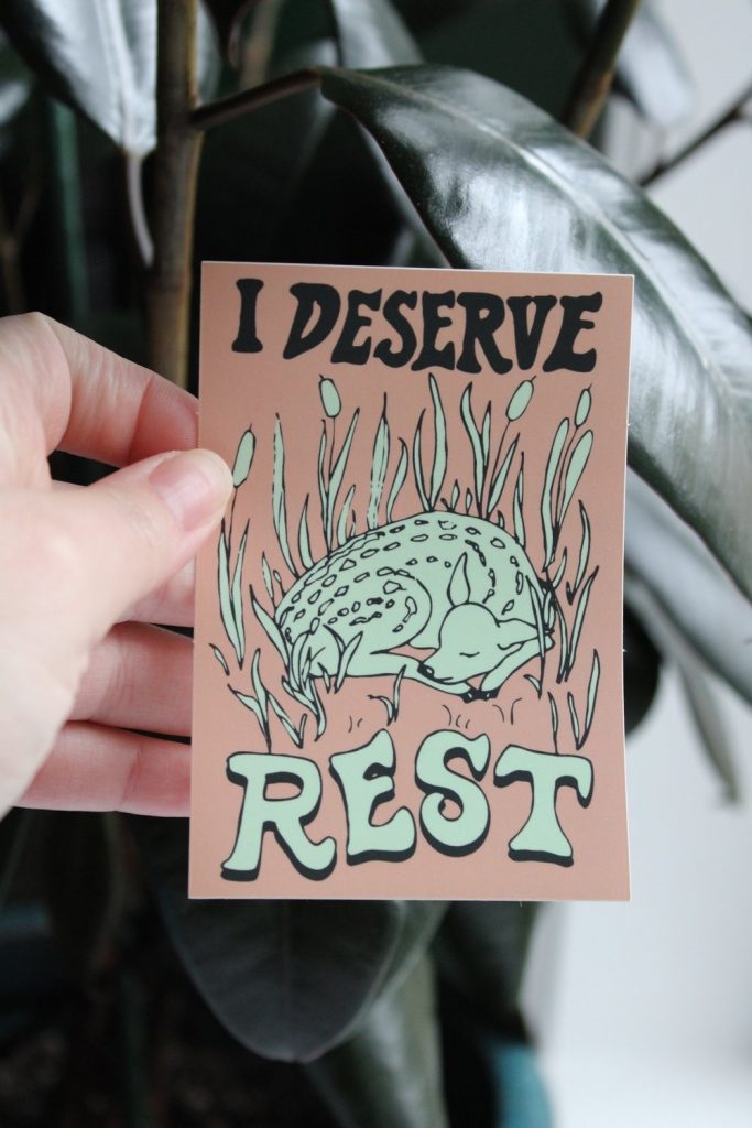 A hand holding a large rectangle sticker. The Text above reads "I deserve" and the text below reads "Rest". In the center is an illustrated baby deer laying in light turquoise grass.