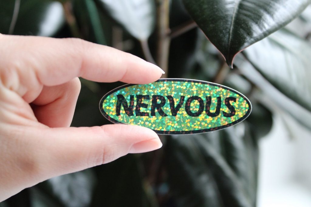 Two fingers holding up an oval sticker. The text in the center reads "Nervous" and the background is a flashy gredn and gold.