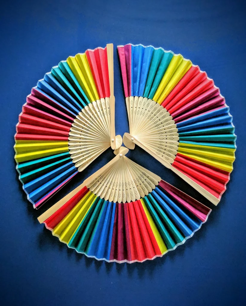 Three rainbow hand held silk fans are laid flat on top of a blue background creating a circle of rainbows.