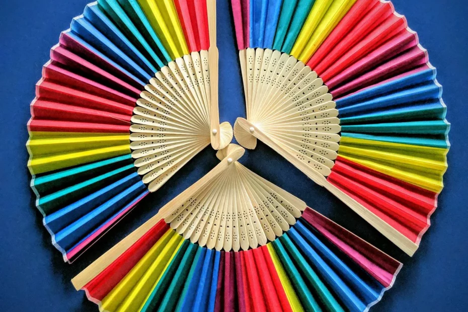 Three rainbow hand held silk fans are laid flat on top of a blue background creating a circle of rainbows. A great pride gift.