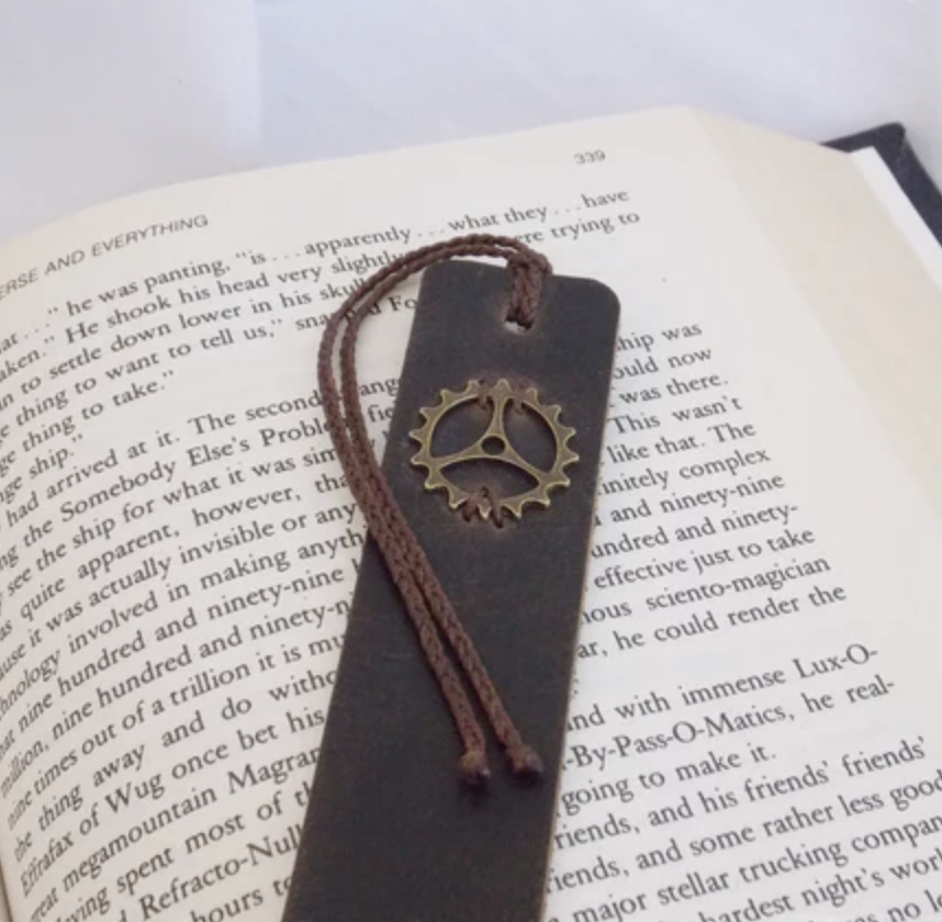 Leather bookmark with metal gear stitched in and long string tassle, placed on top of an open book. 