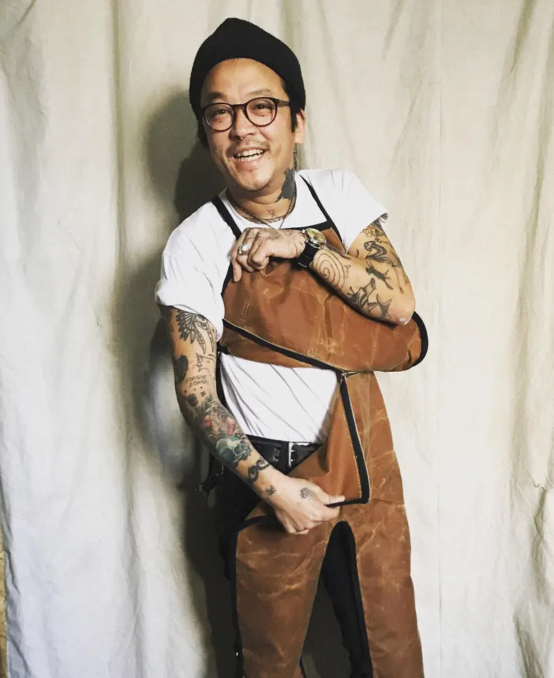 A person is wearing a waxed canvas apron and is displaying how the top bib can unzip from the bottom by lifting apart the two layers.