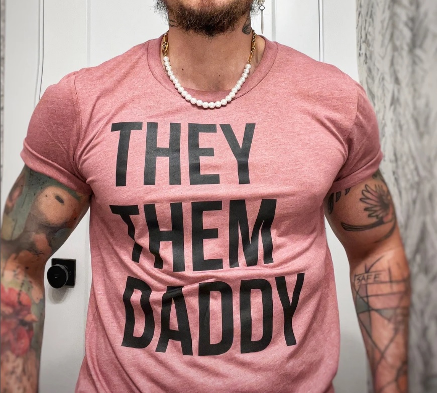 A warm pink t-shirt on a masculine body reads "They Them Daddy" in capitol letters