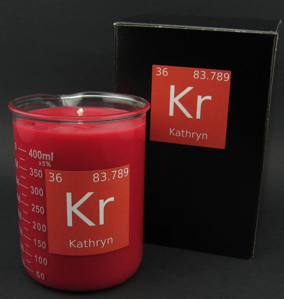 Pink candle in a beaker with a rectangular label that looks like a periodic table entry but reads "Kathryn KR" sits in front of a slightly larger box. 
