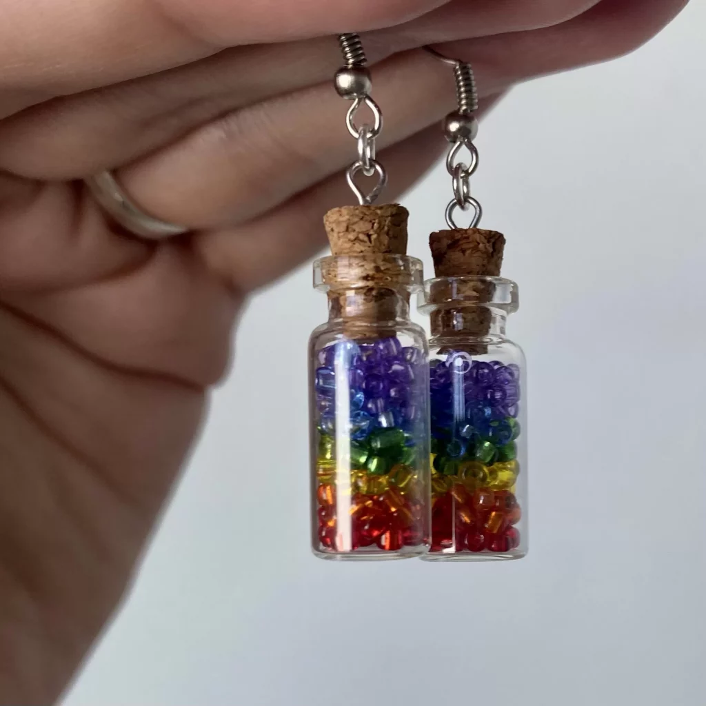 A hand holding up a set of jar earrings filled with beads layered in the colors of the rainbow, starting with purple at the top and ending with read at the bottom. 
