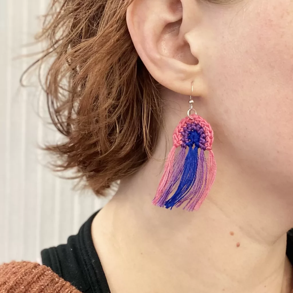 A closeup photo of an earring in someone's ear. The earring made of thin yarn crocheted in the shape of the rainbow, but with colors of the bisexual flag (pink, blue and purple). The rainbow ends in four tassels. 