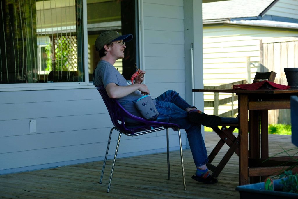 A side photo of Lee sitting on a porch while  knitting. Ze is looking off camera and grinning. 