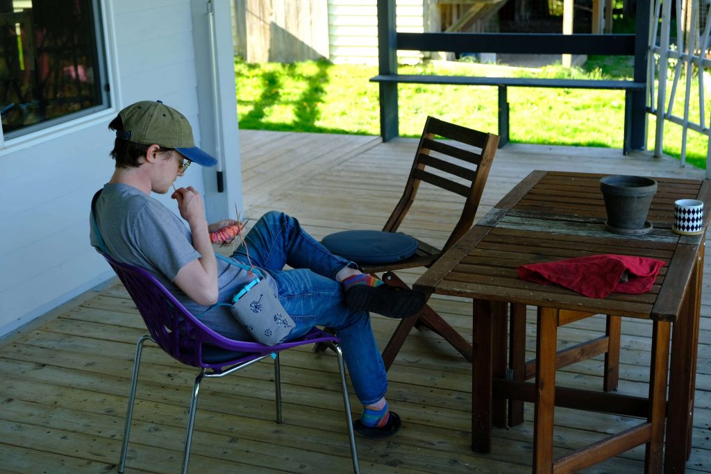An angled photo of Lee sitting on a porch while focusing on hir knitwork.