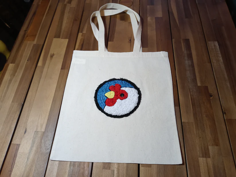 Hand embroidered Artisans Cooperative logo on a white tote bag displayed on a wood floor.