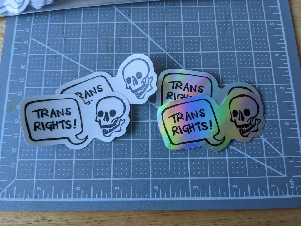 A photo of two sets of stickers, one plain white, and one holographic. They both depict a skull with a speech bubble that says, "TRANS RIGHTS!"