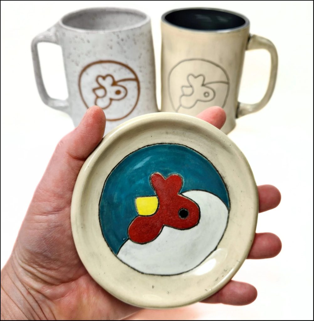 A hand holds the artisan co-op logo glazed on a tea saucer with two mugs in the background containing the same chicken logo.