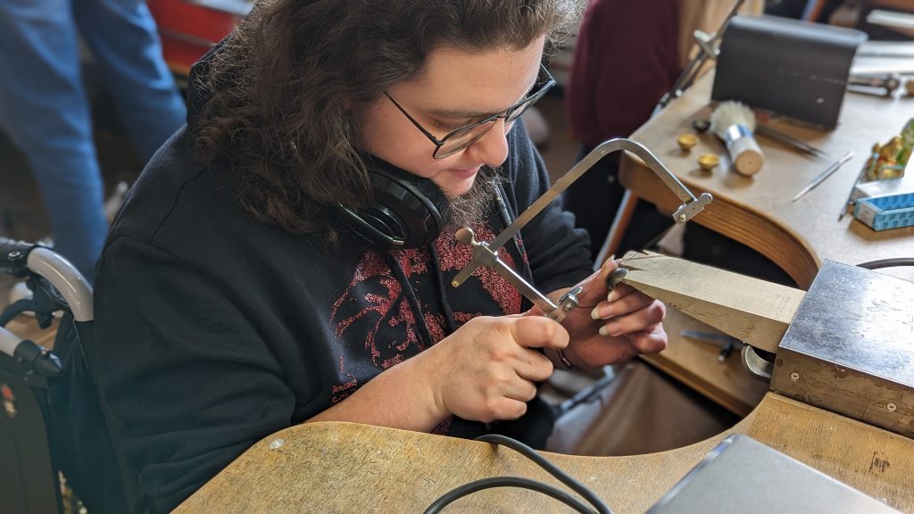 A photo of Tyler sitting in a wheelchair at a workbench using a jewelry saw. They have shoulder-length brown hair and are wearing glasses, and a black hoodie and black pair of headphones resting on their neck. 
